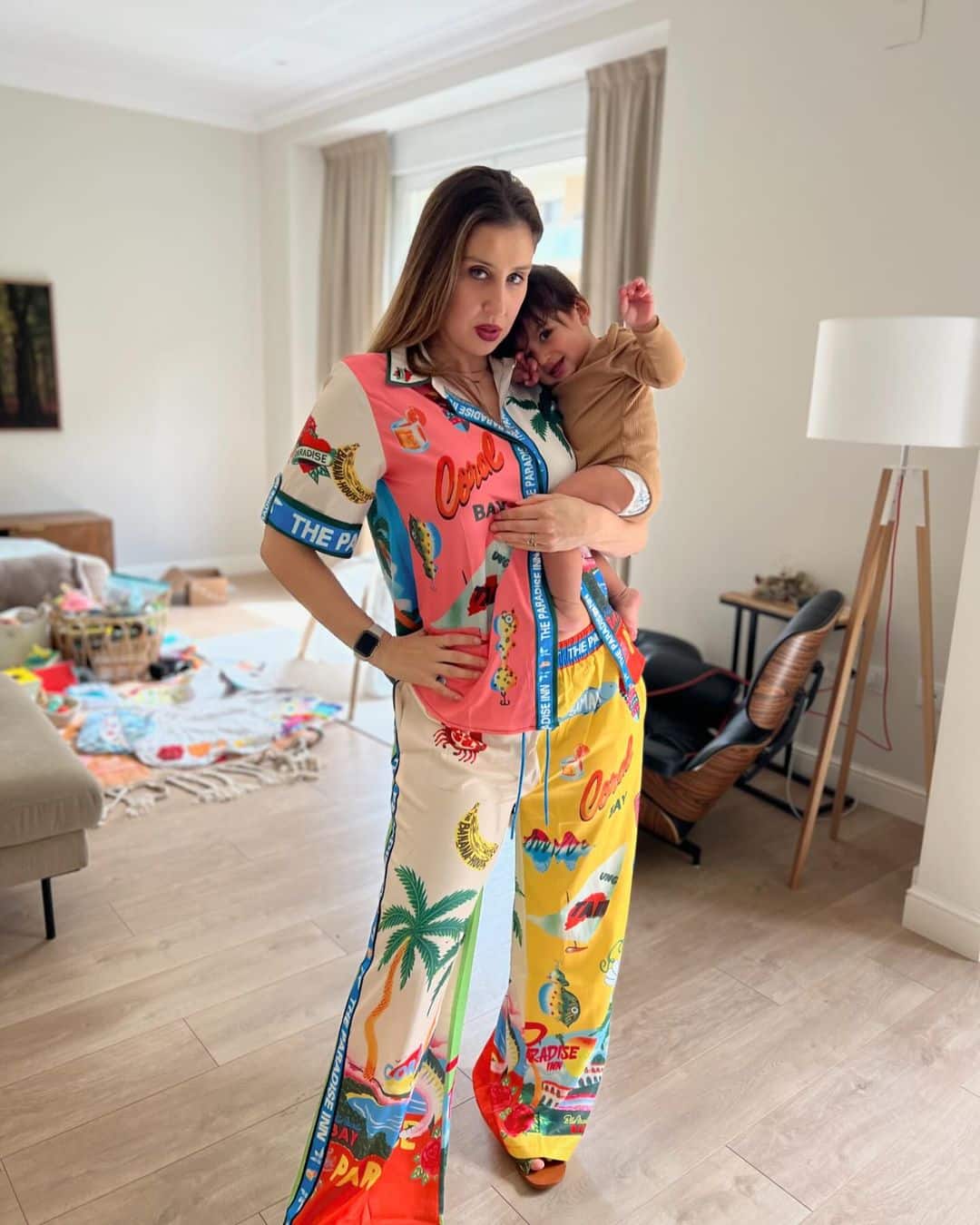 Elise standing in her living room, striking a pose with her hand on her hip and holding her baby in her other arm. She is wearing a brightly colored multi patterned outfit.
